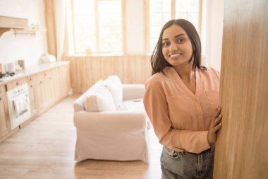 Genç Hindu kadın, güneşli bir evde, ahşap bir panele yaslanmış duruyor. Parlak gülüşü, rahat yaşam alanının sıcak atmosferini tamamlıyor.