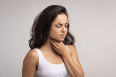 Mutsuz, genç esmer, gözleri kapalı, boğazı ağrıyan beyaz bluz giyen, boynundaki iltihaplı bölgeye dokunan, boş bir yer, gri stüdyo arka planı olan bir kadın.