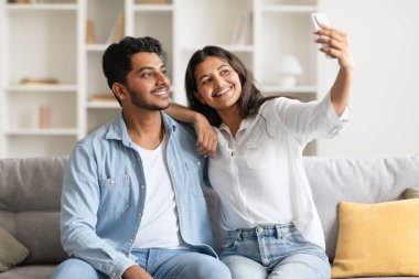 Genç, neşeli Hintli çift koltukta rahatça oturuyor ve samimi anları selfie ile yakalıyorlar. İçten gülümsemeleri, ortak mutluluklarını ve bağlarını yansıtıyor..