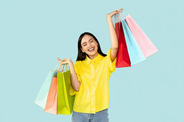 Happy customer. Emotional young brunette woman holding bright shopping bags and smiling, raising hands up, looking at camera over blue background, copy space