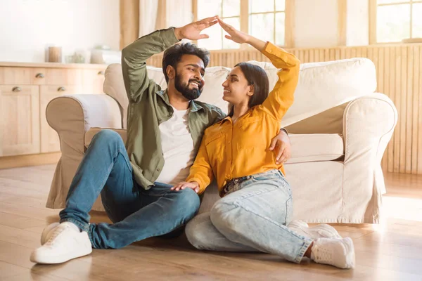 Joyful young couple sitting together on floor near couch in bright room, playfully creating house shape with their hands. They share moment of happiness and laughter in their cozy living space