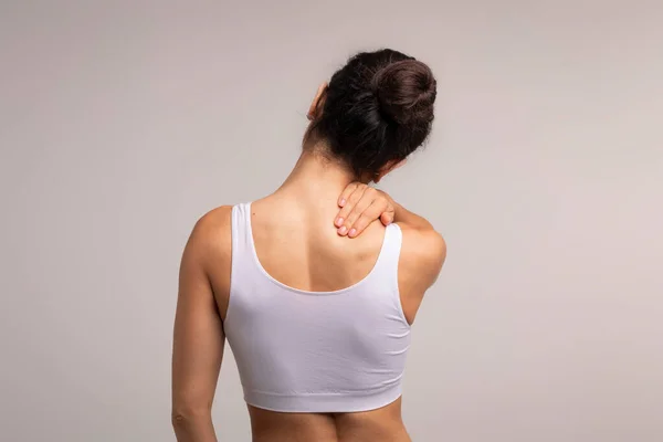 Unrecognizable brunette woman with neck and back pain rubbing her painful body neck area, lady suffering from office syndrome, back view, grey studio background, copy space