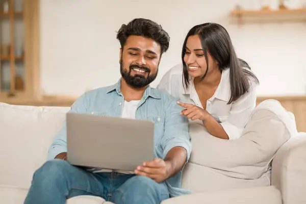 Young couple in casual attire seated on sofa in living room, sharing a moment while browsing on laptop, expressions suggest collaborative interaction and shared interest