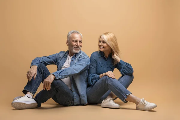 stock image Cheerful beautiful senior couple sitting on floor over beige studio background. Elderly man and woman wearing denim casual outfit have break, rest, chatting and smiling