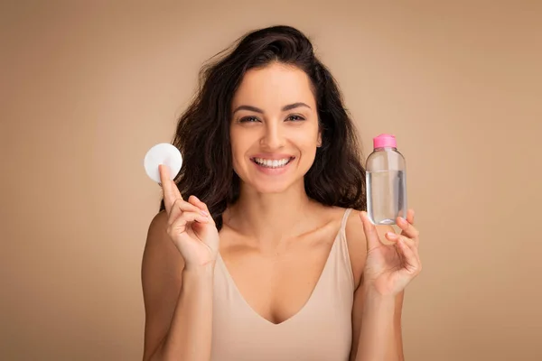Happy smiling pretty naked young eastern brunette woman using cotton pad, cleansing makeup with micellar water, beige studio background, copy space, closeup