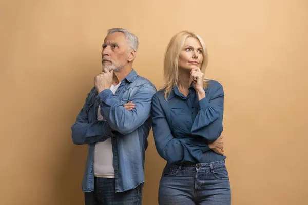 Pensive elderly couple thinking about something, thoughtful serious senior man and woman standing back to back, touching chin and looking aside, looking for solution, beige background