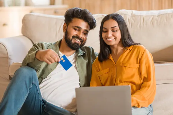 若いインドのカップルはソファに座って ラップトップでブラウジング 男は青いクレジットカードを保持し オンラインショッピングや支払いを示し 彼らは両方コンテンツを見て従事 — ストック写真
