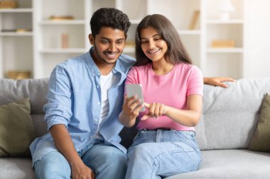 Genç Hintli eşler rahat bir şekilde kanepede oturuyor, kadın cep telefonu ekranına işaret ediyor, partnerlerinin dikkatini çekiyor.