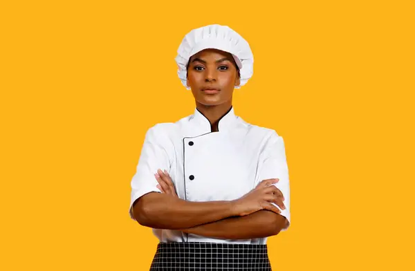 Confident Young Black Chef Woman In Uniform And Hat Posing Isolated Over Yellow Background, Beautiful African American Cook Female Standing With Folded Arms And Looking At Camera, Copy Space