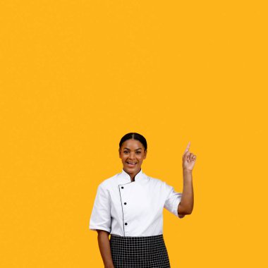 Mutlu Afrikalı Amerikalı kadın aşçı parmaklarıyla fotokopi uzayı işaret ediyor, üniformalı aşçı kadın kafasının üstünde bedava bir yer gösteriyor, sarı stüdyonun arka planında tek başına duruyor.