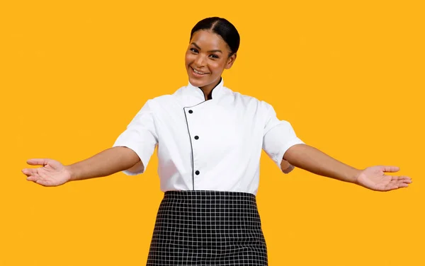 Beautiful black chef woman chef showing empty palms, spreading arms at camera, happy african american cook female presenting invisible objects or gesturing welcome while posing on yellow background