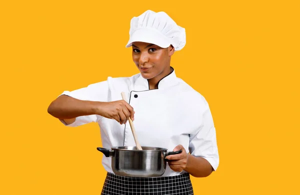 Cooking Concept. Smiling Black Chef Woman Stirring Food In Saucepan While Standing Isolated Over Yellow Background, Young African American Cook Lady In Uniform Using Wooden Spatula, Preparing Meal