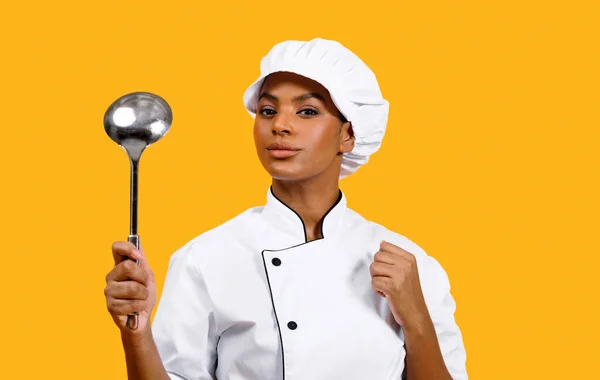 Confident Young Black Chef Woman In Uniform Holding Metal Ladle In Hand And Looking At Camera, Beautiful African American Cook Lady Standing Isolated Over Yellow Studio Background, Free Space