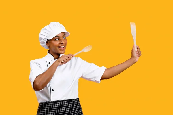 Playful Young Black Chef Lady With Spatulas In Hands Posing On Yellow Studio Background, Happy African American Cook Woman Wearing Uniform Having Fun With Kitchen Utencils, Copy Space