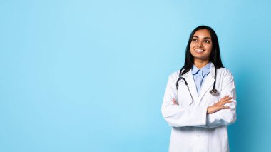 Orta Doğulu kendine güvenen genç doktor el ele tutuşuyor, mavi arka planda tıbbi önlükle gülümsüyor, sağlık reklamları için sağlık sektöründen profesyonel doktor çekimleri yapıyor. Panorama, boşluk
