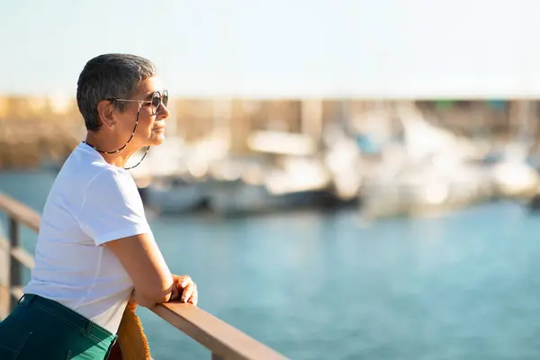Summer Moments. Side view of mature lady looking at sea with vessels in the backdrop, standing at pier outside. Seaside break for seniors. Blank space for vacation travel offer ad