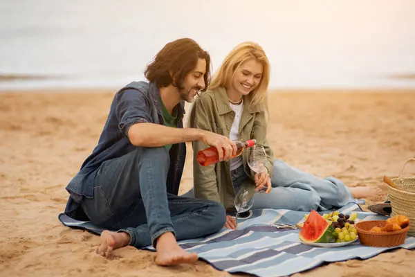 Affectionate Young Couple Having Romantic Picnic On The Beach, Happy Millennial Man And Woman Drinking Wine And Having Fun Together, Enjoying Date Outside, Celebrating Anniversary, Free Space