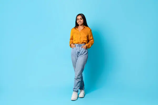 Confident Indian lady standing full length in casual jeans with hands in pockets, posing against blue studio background, perfect for ads or promotions. Free space for text. Modern fashion and style