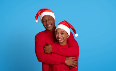 Afro-Amerikalı çift, mavi stüdyo arka planlarına sıcak bir şekilde sarılıyor, Noel bayramı neşesi ve sevgi dolu bir bağ sergiliyor, kırmızı kazak ve Noel Baba şapkası giyip poz veriyorlar.