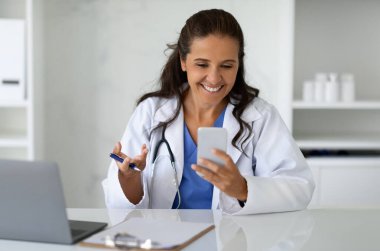 Uzaklık sağlık hizmetleri, tele sağlık teknolojileri. Dolabında dizüstü bilgisayar, cep telefonu ve fotokopi makinesi kullanarak oturan mutlu olgun kadın doktor. Klinikte çalışan kadın doktor.