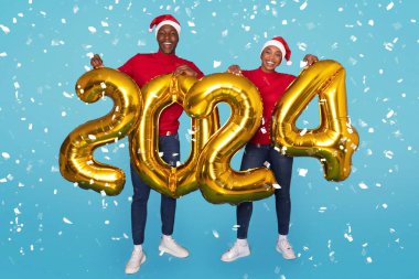 Altın 2024 Balon Duran Afro-Amerikan Çifti Turkuaz Mavi Arka plan, Düşen Konfeti 'nin ortasında Yeni Yılı Kutluyor, Noel Şapka Giyiyorlar. Tam Uzunluk Çekimi