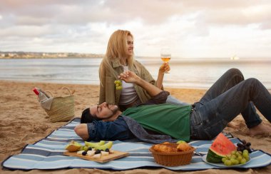 Tutkulu genç bir çiftin portresi sahilde pikniğin keyfini çıkarıyor, romantik bir milenyum erkeği ve kadın şarap içiyor ve yan yana gevşiyor, ekose üzerinde dinleniyor ve dışarıda eğleniyor.