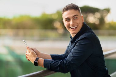 Günlük hayattan zevk alan gülümseyen genç Avrupalı adam, telefonda yazıyor, şehirde ve dışarıda boş alana bakıyor. İş, iş, cihaz ile çalışma, serbest çalışma, sosyal ağlarda sohbet, iyi haber