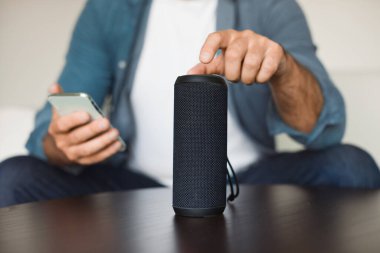 Kablosuz akıllı hoparlör ve modern akıllı telefon vasıtasıyla evde müzik aletleri ve uygulamalar kullanarak müzik dinleyen tanınmayan bir adam. Telefonlu ve müzikal kolonlu bir adamın kesilmiş fotoğrafı.