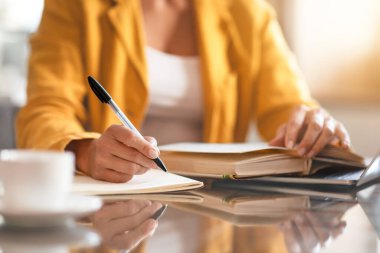 Sarı ceketli bir kadının not defterine kalemle yazması, masadaki bir fincan kahvenin yanında iş kadını evde otururken not alması. Kırpılmış atış