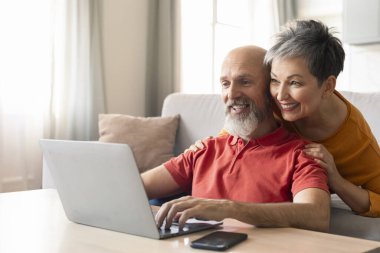 Evde dizüstü bilgisayar kullanan mutlu yaşlı çiftin portresi, oturma odasında bilgisayarla dinlenen yaşlı eşler, internetten alışveriş yapan veya birlikte fotoğraf izleyen eşler,
