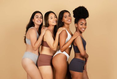 Four happy and diverse women pose together, radiantly smiling, showcasing a range of stylish underwear against neutral beige studio background clipart