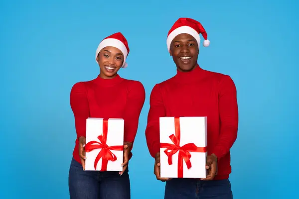 Joyful Black Spouses Presenting Wrapped Xmas Gift in Santa Hats, Posing Holding Two Presents Boxes with Delight on Blue Studio Background, Celebrating Winter Holidays and Sharing New Year Offers