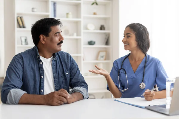 Indian Female Doctor In Blue Uniform Consulting Middle Aged Male Patient During Appointment Meeting In Clinic, Friendly Therapist Woman Talking To Mature Man, Explaining Medical Therapy Process