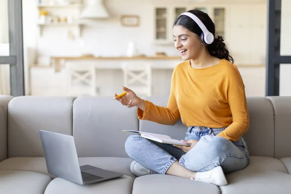 Online Tutoring. Smiling Indian Woman In Headphones Making Teleconference With Laptop, Young Eastern Female Teacher Talking And Gesturing At Web Camera During Virtual Lesson, Sitting On Couch At Home