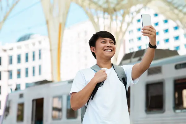 Cheerful excited asian guy with backpack travel blogger taking selfie by phone, recording video for followers while standing on platform at train station, copy space. Tourism