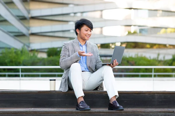 Young Asian businessman conducting an online meeting via laptop, sitting outdoors in urban city area. Guy in suit talking to computer webcam video calling. Modern corporate communication