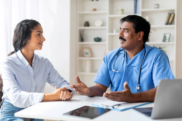 Smiling Indian Doctor Man Talking To Female Patient During Appointment In Office, Professional Male Therapist In Blue Scrubs Uniform Consulting Young Woman, Explaining Medical Treatment Process