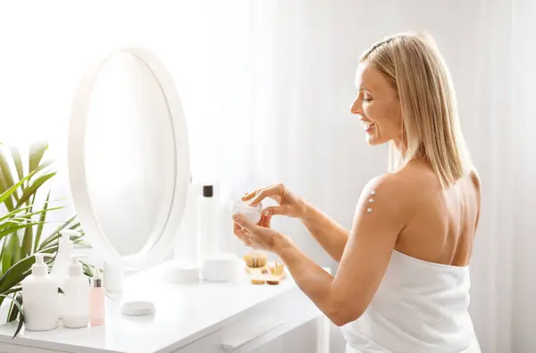Beauty Care Concept. Smiling Middle Aged Woman Opening Jar With Moisturising Cream While Sitting At Dressing Table At Home, Happy Mature Female Wrapped In bath Towel Making Skincare Routine