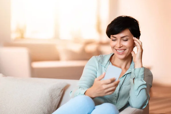 Happy middle aged European woman scrolling on smartphone, enjoying casual chat, sitting on couch in living room indoor. Domestic comfort meets digital communication concept. Free space