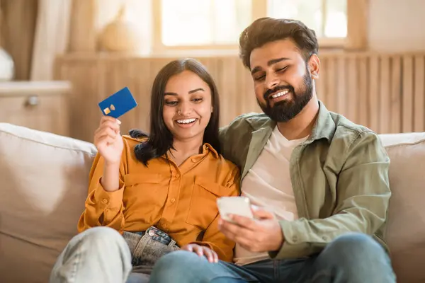 Joyful Indian couple seated on sofa at home, woman holding blue credit card while man smiles, holding smartphone. Captures moment of online shopping or financial discussion