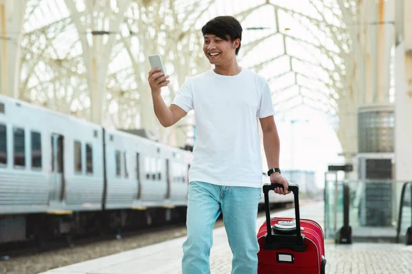 Chinese millennial guy in casual outfit tourist walking by train station, using smartphone, carrying luggage. Traveler using traveling mobile application, checking train schedule online