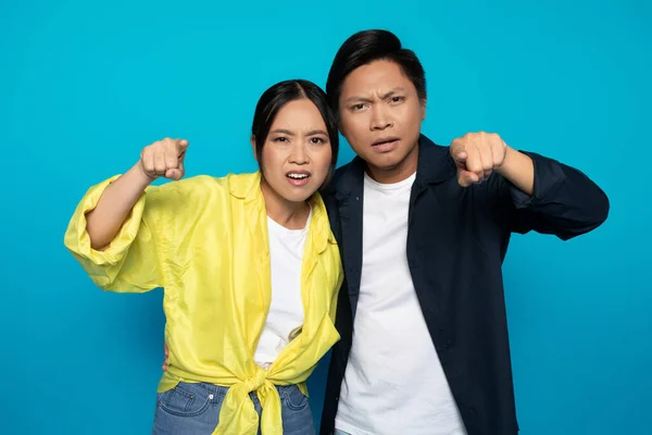 Confused sad serious asian couple in casual point fingers at camera, isolated on blue studio background. Emotions gesture, what is this, expression and question, free space