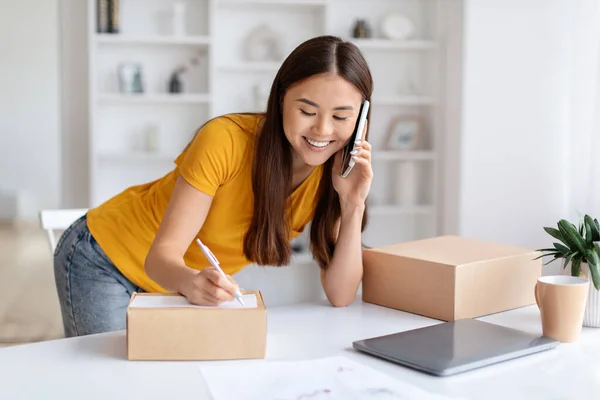 笑顔のアジアの女性は 自宅のオフィスで働いている間 携帯電話を介して注文を受け取ります 配送のための小包箱に書く若いビジネスマン 顧客との携帯電話で話す — ストック写真