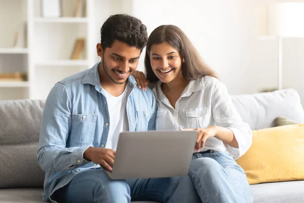 Young Indian couple enjoys leisurely moment at their homey living room, sharing laptop and exploring content together, their expressions filled with interest and delight