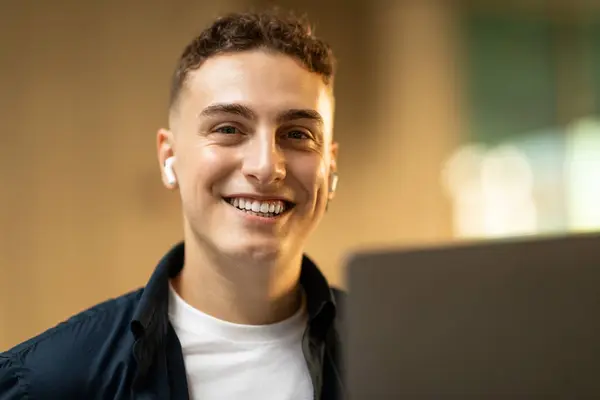 Smiling attractive young european guy in casual, wireless headphones enjoy lifestyle, work on laptop in city cafe, office. Work, successful business, study, education with device, freelance