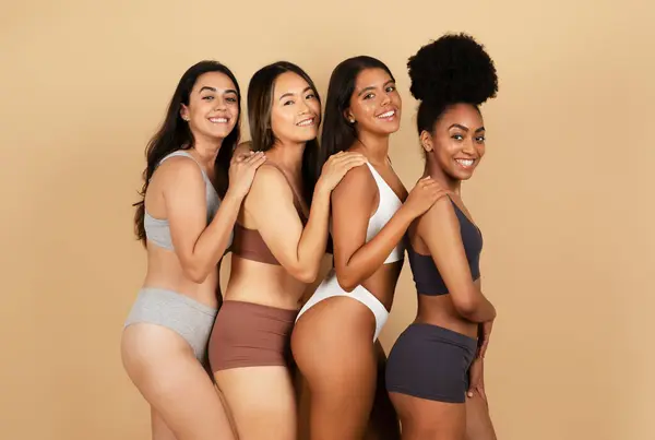 Four happy and diverse women pose together, radiantly smiling, showcasing a range of stylish underwear against neutral beige studio background