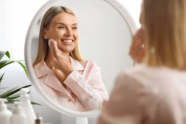 Smiling Beautiful Mature Woman Cleansing Skin With Cotton Pad While Making Daily Beauty Routine At Home, Attractive Middle Aged Lady Enjoying Skincare Treatments, Looking In Mirror At Her Reflection