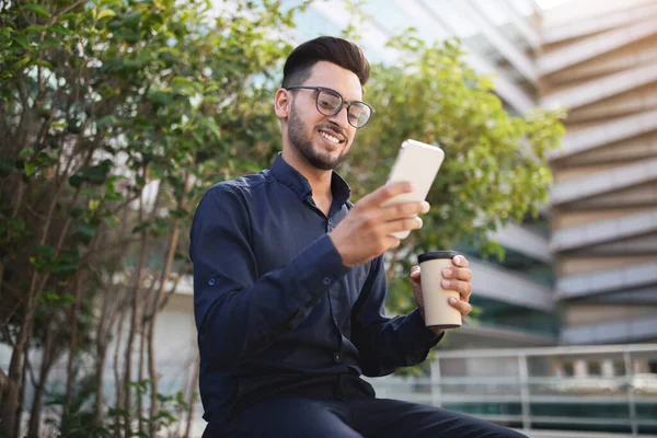 Millennial Arabian business guy holds smartphone immersed in websurfing on mobile device, enjoying coffee break sitting in urban area outdoors, wearing eyewear. Internet and communication