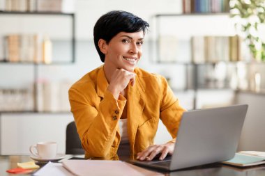 Orta yaşlı Avrupalı iş kadını, kendine güvenen bir gülümsemeyle dizüstü bilgisayarda çalışıyor, kapalı ofis masasında oturuyor. İş dünyasında başarılı kadınlar.