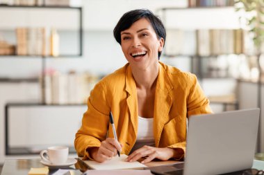 Orta yaşlı mutlu esmer iş kadını ofis masasında dizüstü bilgisayarla kağıt yazıyor. Belgeler ve iş sözleşmeleri ile çalışan başarılı şirket yöneticisi notlar alıyor